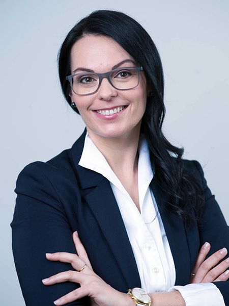 Porträt von Monika Valcanover mit vor der Brust verschränkten Armen, sie trägt einen dunkelblauen Blazer über einer weißen Bluse. Sie lächelt mit geöffneten Mund und trägt eine große, dunkle Brille.