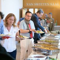 Nach den interessanten Vorträgen gab es Stärkung am Buffet. Zahlreiche Gäste stehen mit teilweise beladenen Tellern vor den geöffneten Essensbehältern und bedienen sich selbst.