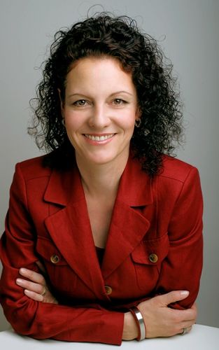 Susanne Schwanzer trägt einen roten Blazer, sie stützt ihren Oberkörpfer auf die Tischplatte vor sich und lächelt freundlich in die Kamera.