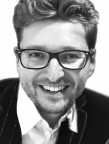 Andreas Lederer trägt eine eckige Brille und lächelt mit geöffneten Mund in die Kamera. Er trägt ein weißes Hemd unter einem schwarzen Sakko. Das Bild ist Schwarz-Weiß.