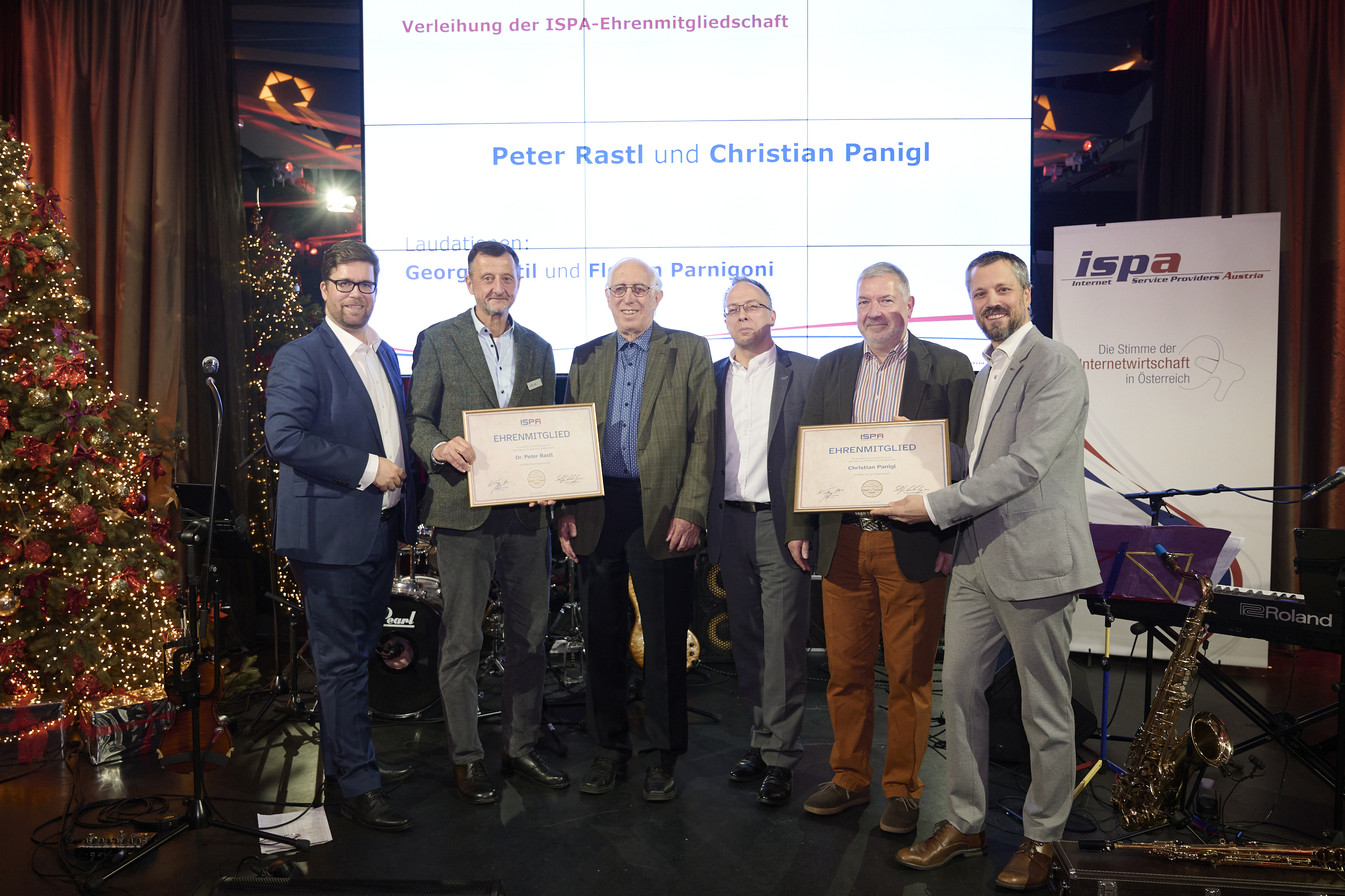 Die &Uuml;berreichung der ISPA-Ehrenmitgliedschaften, auf der B&uuml;hne stehen vor einem Weihnachtsbaum und einem ISPA-Roll-up von links nach rechts: Stefan Ebenberger (ISPA-Generalsekret&auml;r), Georg Chytil (Laudatio f&uuml;r Peter Rastl), Peter Rastl, Harald Kapper (ISPA-Pr&auml;sident), Christian Panigl, Florian Parnigoni (Laudatio f&uuml;r Christian Panigl)