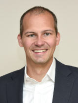 Nico Grove Portrait von Nico Grove, er trägt ein weißes Hemd unter einem schwarzen Sakko. Er lächelt freundlich mit geöffneten Mund in die Kamera, seine Zähne sind zu sehen.