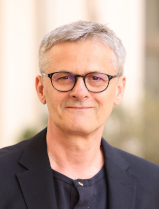 Alois Schrems blickt mit geschlossenem Mund l&auml;chelnd in die Kamera. Er tr&auml;gt eine Brille mit gro&szlig;em, dunklem Rahmen, ein schwarzes Sakko &uuml;ber einem schwarzen Leiberl.