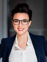 Christine Antlanger-Winter l&auml;chelt freundlich mit offenem Mund in die Kamera. Sie tr&auml;gt eine wei&szlig;e Bluse unter einem dunkelblauem Blazer und eine gro&szlig;e Brille.